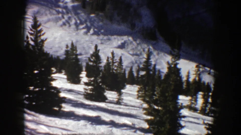 1961 Aspen frame showing a bright snow slope with clustered evergreen trees and shadowed mountain terrain in the background.