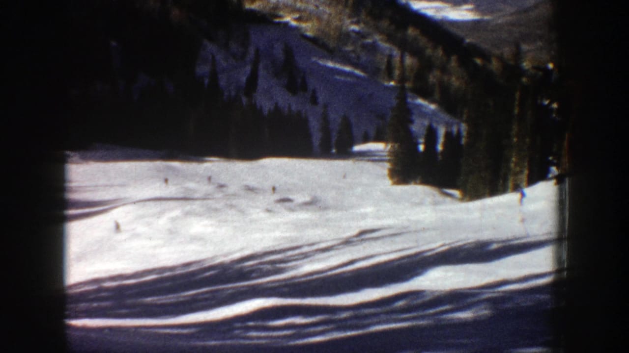 Open snowfield below wooded mountain slopes with tiny distant skiers.
