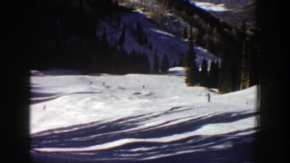 Aspen, Colorado 1961 home-movie frame showing scattered skiers on a broad snowy run bordered by dark evergreens.