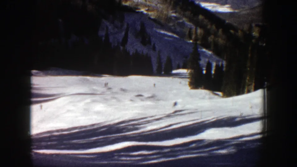 Aspen, Colorado 1961 frame of a sunlit ski slope with distant skiers, evergreen trees, mountain terrain, and dark 8mm edge shading.