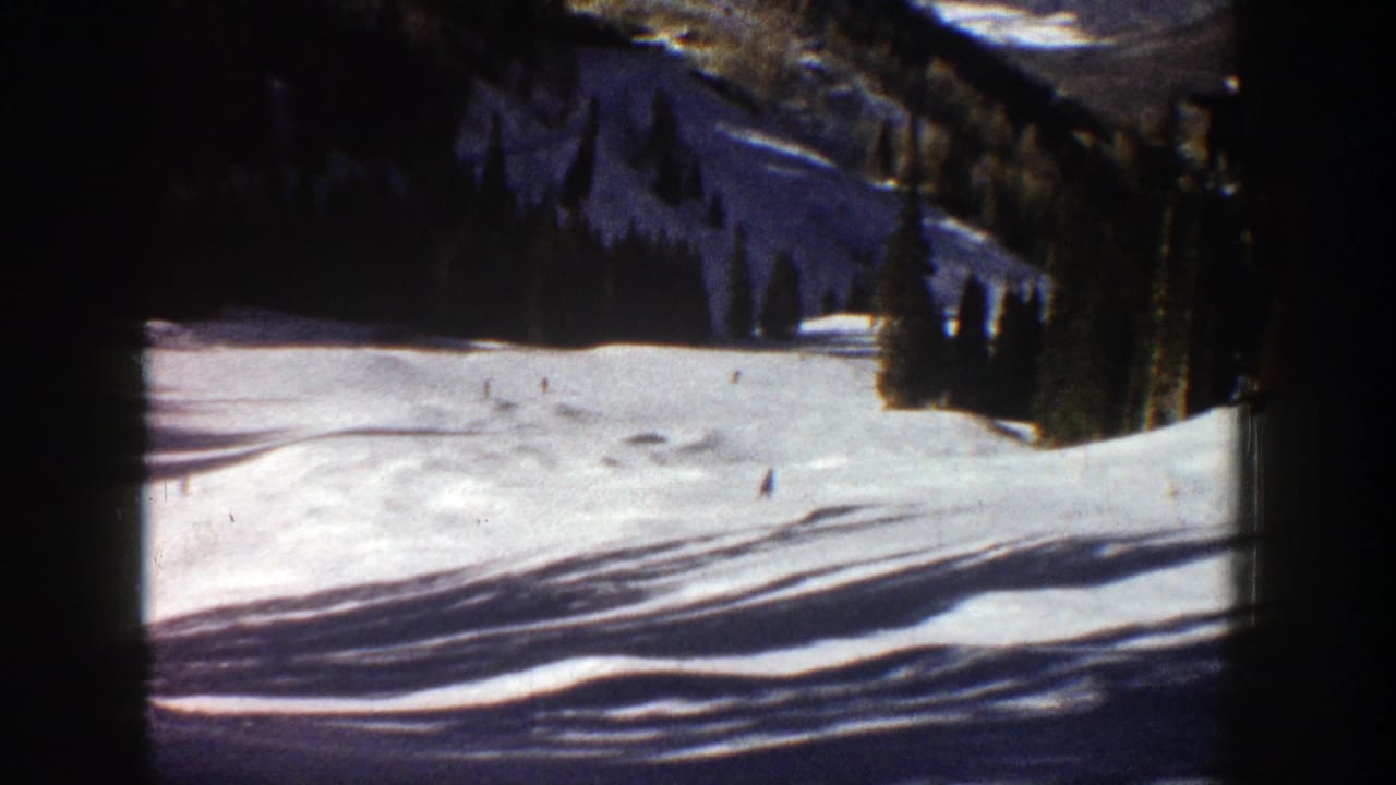 1961 Aspen home-movie frame showing a wide snow-covered ski run with tiny skiers below evergreen trees and mountain terrain.