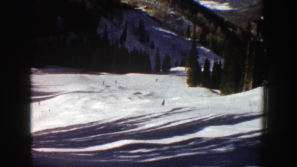 1961 Aspen home-movie frame showing a wide snow-covered ski run with tiny skiers below evergreen trees and mountain terrain.