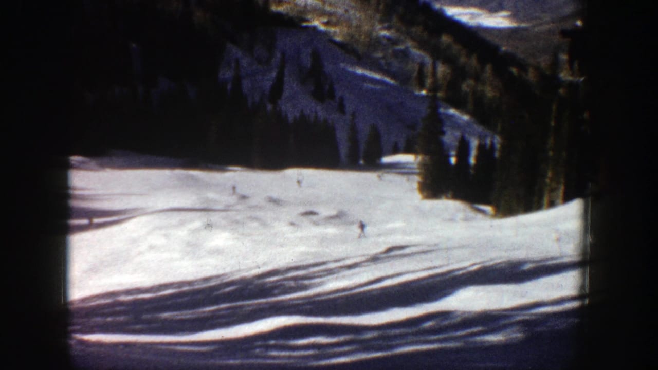 Snowy mountain run with small skiers, tree line, shadows across snow, and visible film edges.