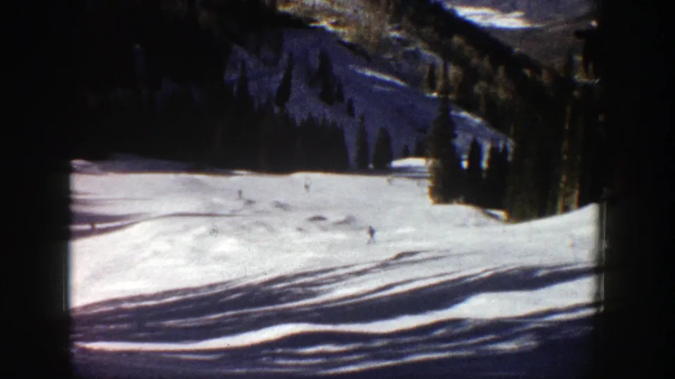 1961 Aspen ski footage frame with small figures descending a bright snowy run beneath dark evergreen stands.