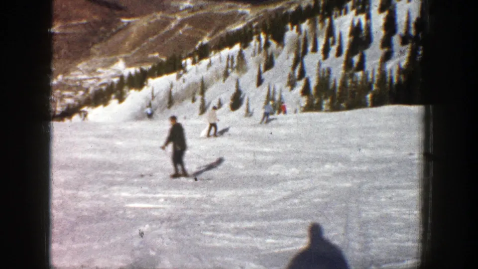 1961 Aspen frame showing skiers spaced across a downhill snowfield, with evergreens and mountain terrain in the background.