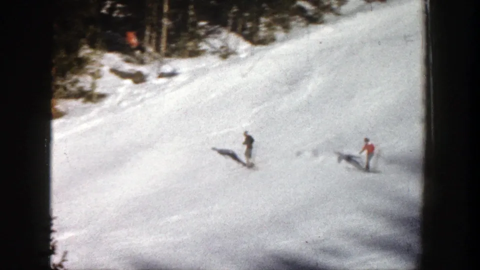 1961 Aspen home-movie frame showing two distant skiers moving downhill near an evergreen-lined section of slope.