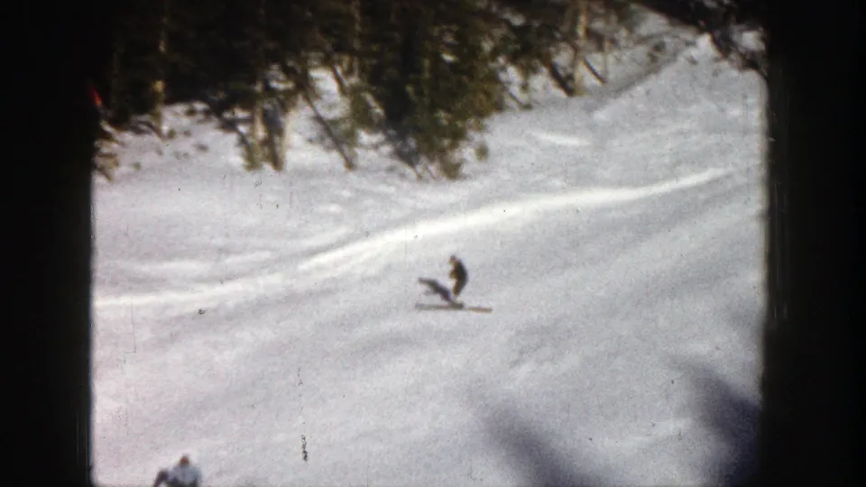 A distant skier crosses a snowy Aspen slope in 1961 home-movie footage, with evergreen trees and visible ski marks behind.