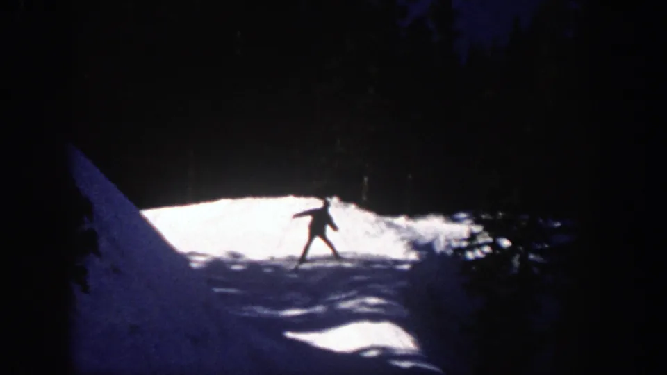 A lone skier crosses a sunlit section of the Aspen slope, partly silhouetted by the dark forest background.