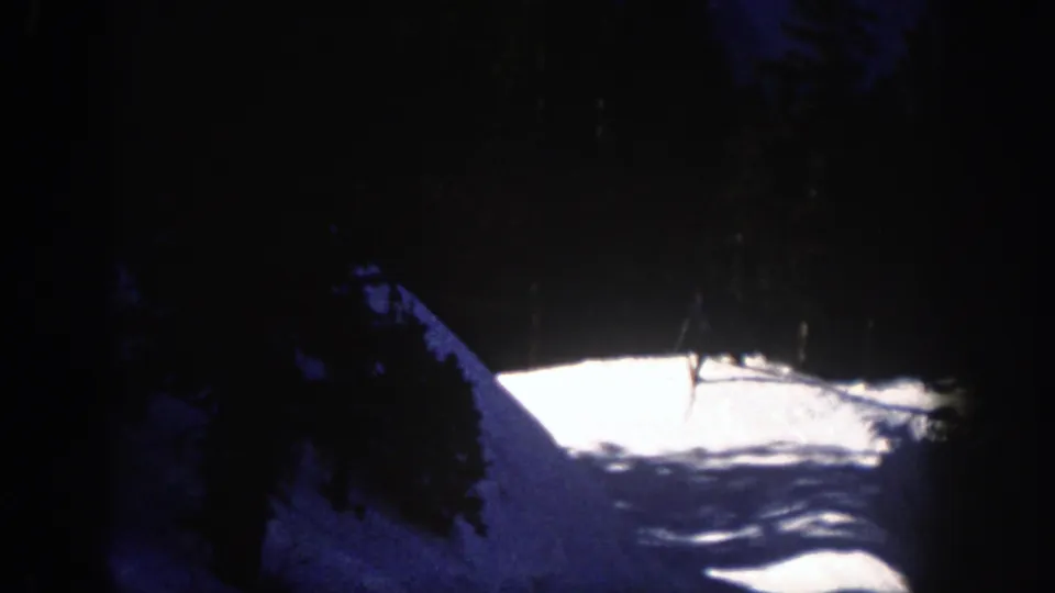 The skier appears near the upper edge of the snowy run, partly obscured by contrast and tree shadows in the 8mm frame.