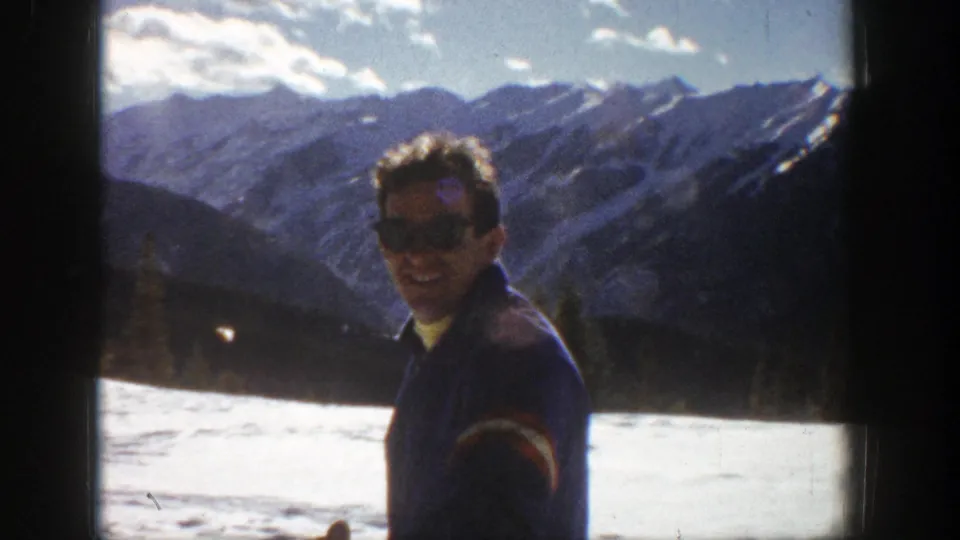 In the 1961 Aspen home movie, the man smiles toward the camera while standing before snow, trees, and distant peaks.