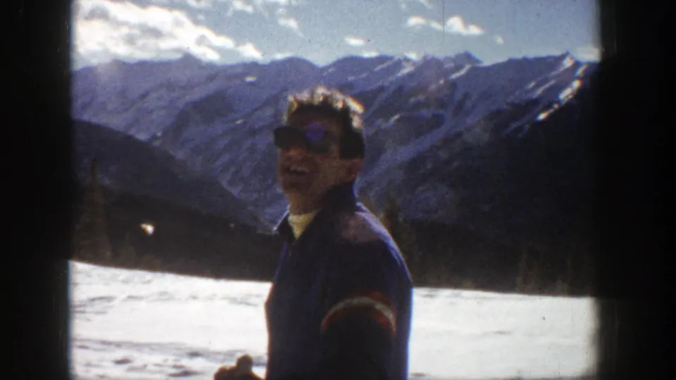 Adult man laughs toward the camera in Aspen, Colorado, with a bright snowfield and ridged mountain backdrop visible behind him.
