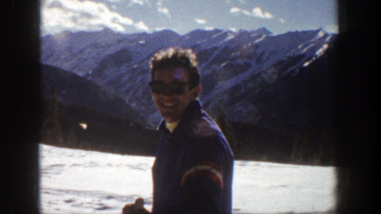 Adult man in dark clothing stands in snowy Aspen, Colorado, with a mountain range behind him in 1961 home-movie footage.