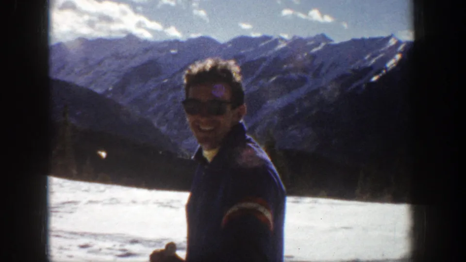 Adult man in dark clothing stands in snowy Aspen, Colorado, with a mountain range behind him in 1961 home-movie footage.