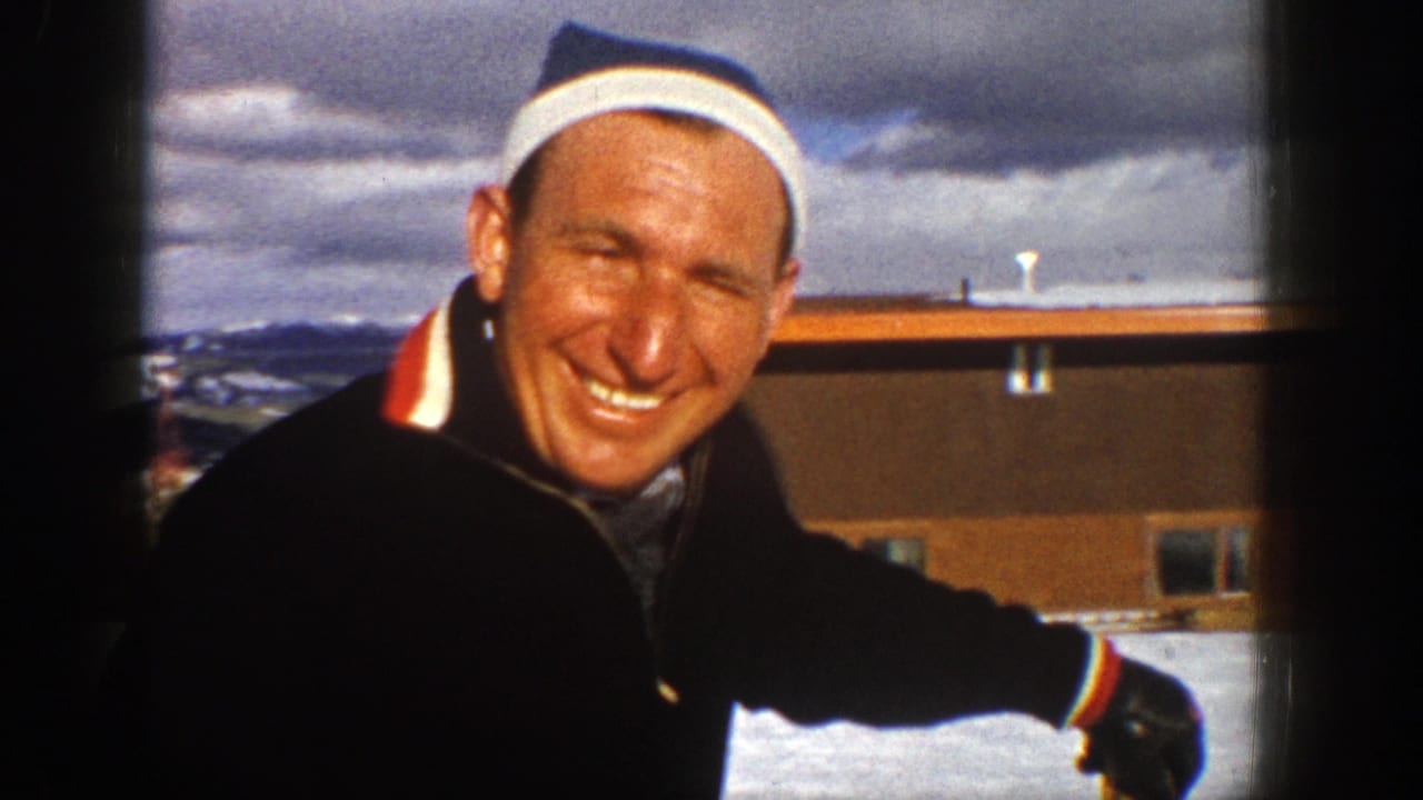 Smiling man in striped headband beside a snowy building and mountains.