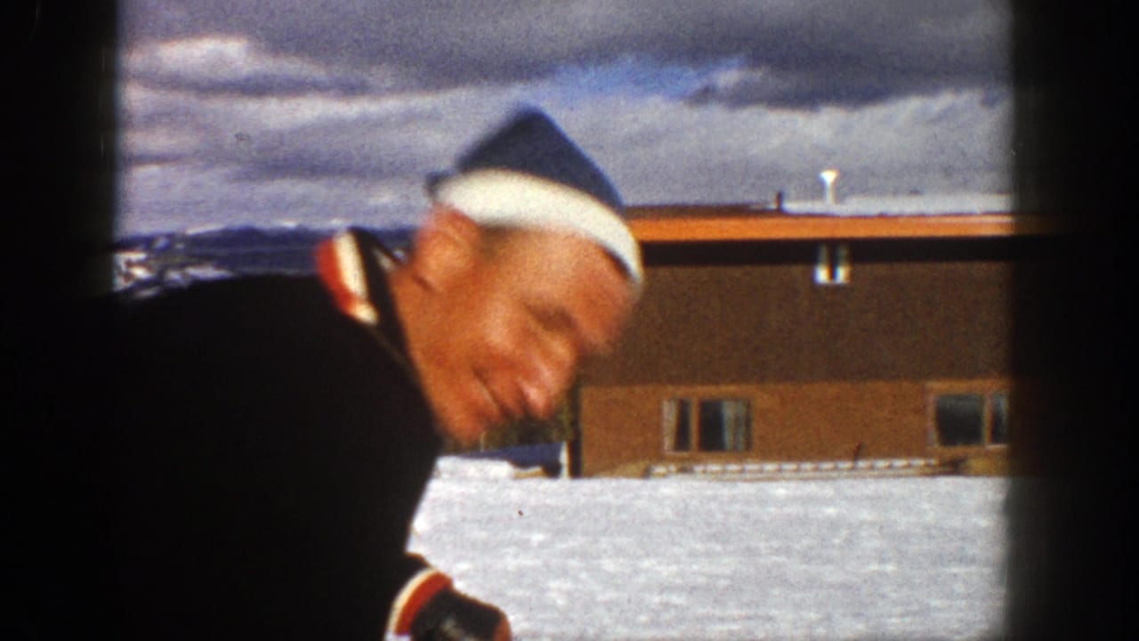 In 1961 Aspen home-movie footage, a man in a winter hat and dark coat leans toward the camera from a ski lift near snow and a low building.