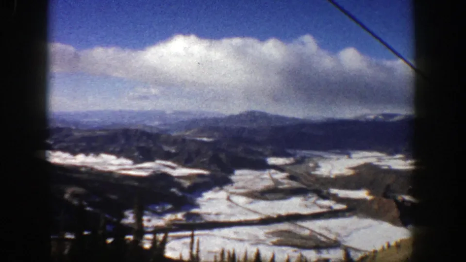 A wide 1961 Aspen mountain view shows snowfields, tree lines, distant peaks, bright clouds, and a diagonal ski-lift cable.