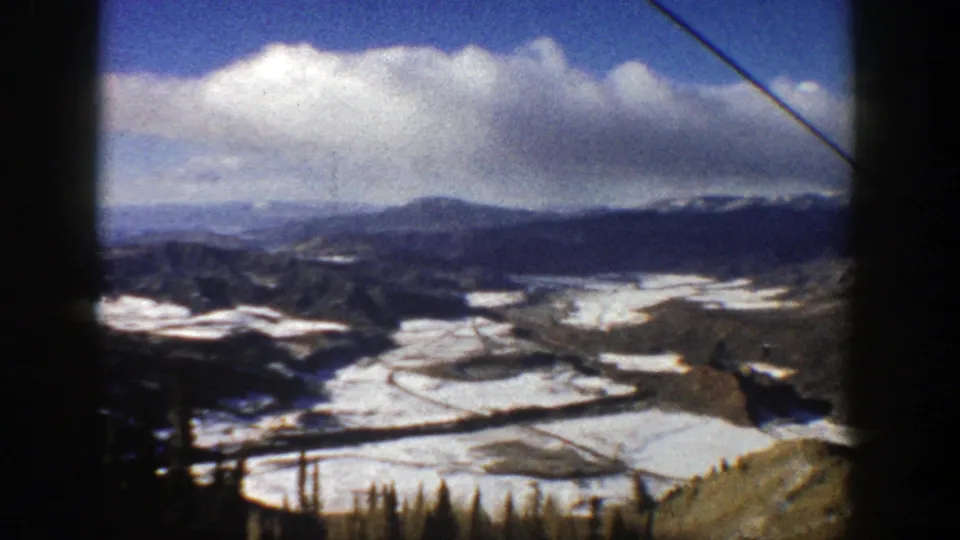 1961 Aspen, Colorado ski-resort view with snow-covered terrain, distant ridges, evergreens, and an overhead lift cable.
