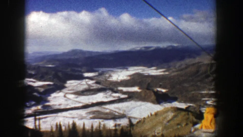 High-angle 1961 Aspen resort panorama showing snowfields, mountains, cloud cover, and a ski-lift cable across the frame.