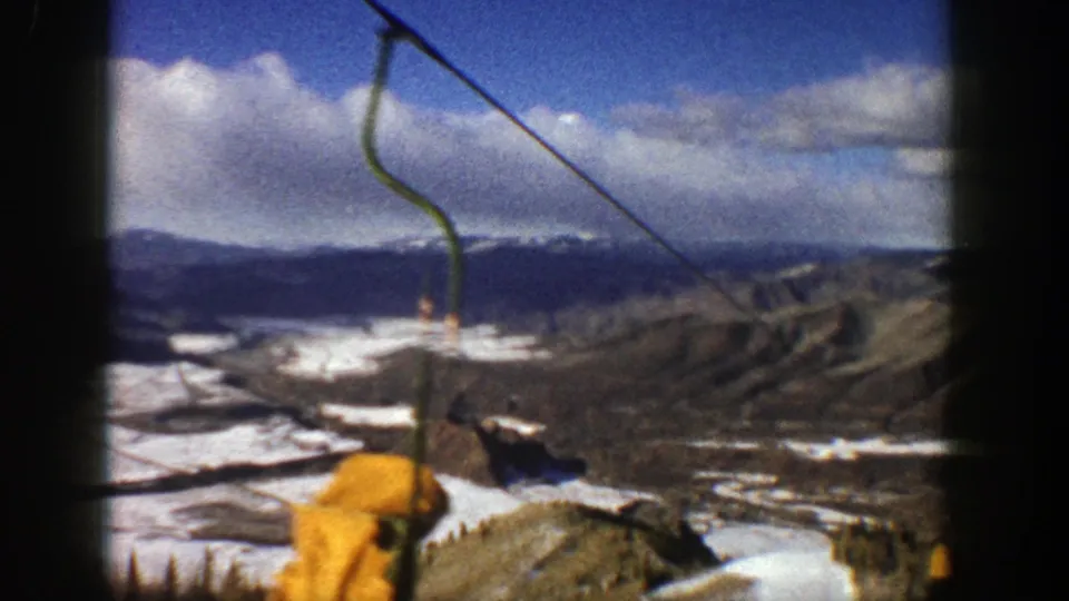 1961 Aspen ski-area view with a yellow cable car entering the foreground above snowy runs and a broad mountain backdrop.