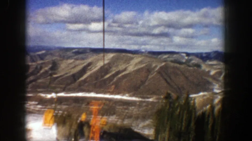 1961 home-movie frame from Aspen showing a snowy slope, evergreen trees, orange lift structures, and distant ridgelines.
