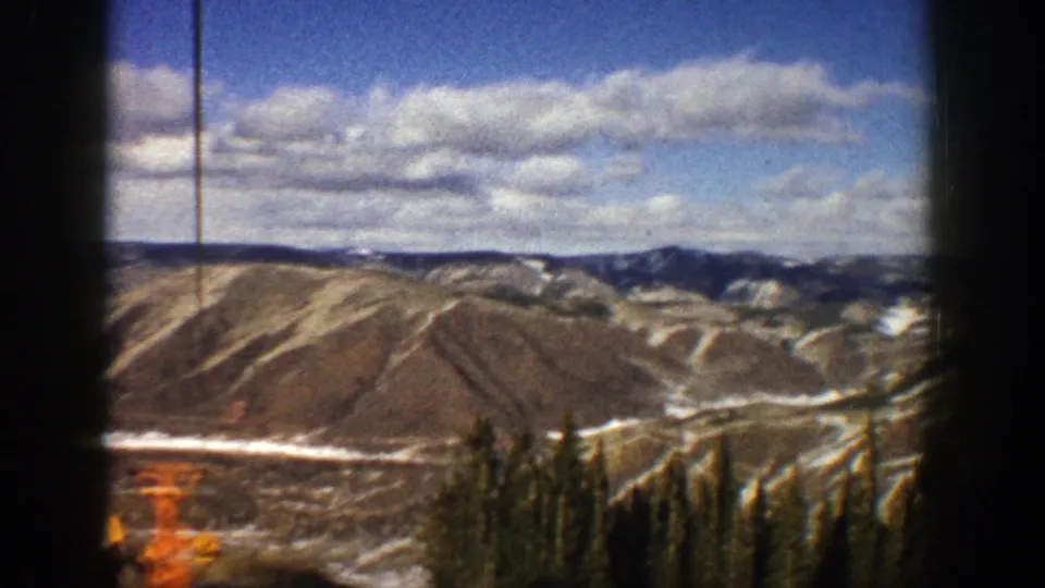Aspen, Colorado mountain scenery from a moving ski lift, with evergreens, orange lift equipment, snow, and 8mm film grain.