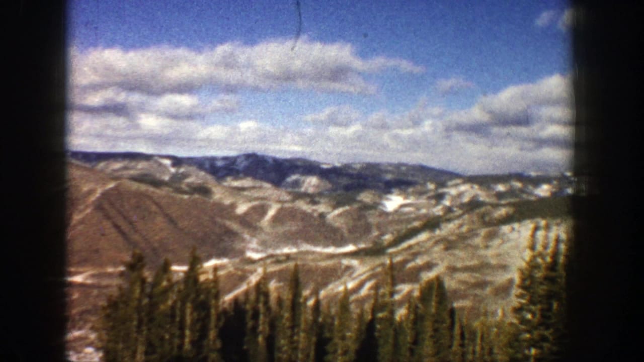 Mountain ridges and patchy snow seen beyond evergreen trees and a dark film edge.