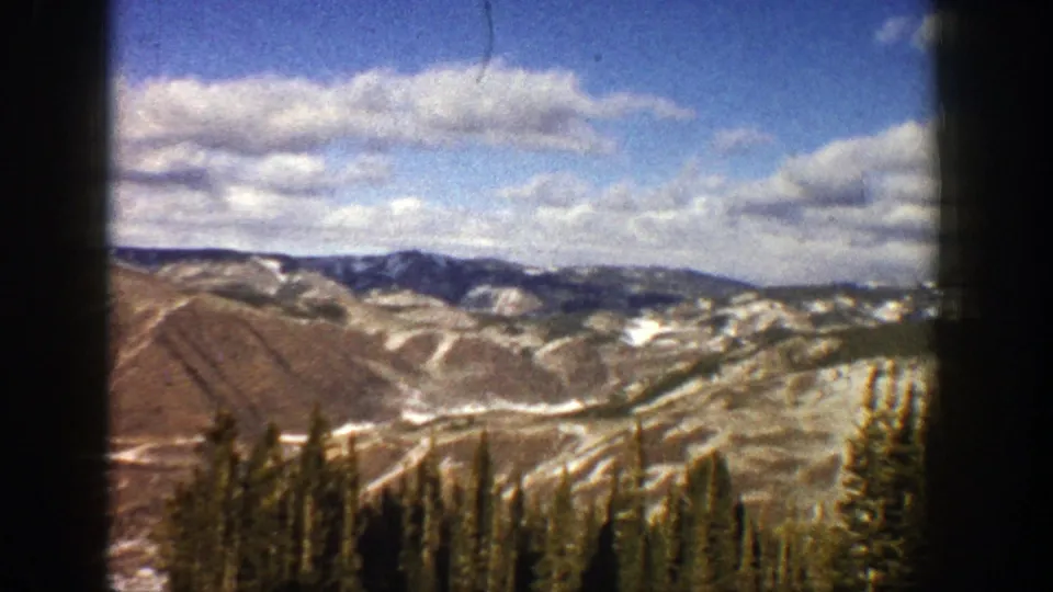 Aspen, Colorado mountain panorama with patchy snow on brown hillsides, evergreen foreground, and cloudy blue sky on 8mm film.