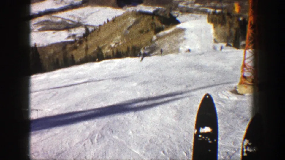 1961 Aspen ski-slope view with foreground skis, packed snow, a lift tower, evergreen trees, and mountain terrain beyond.