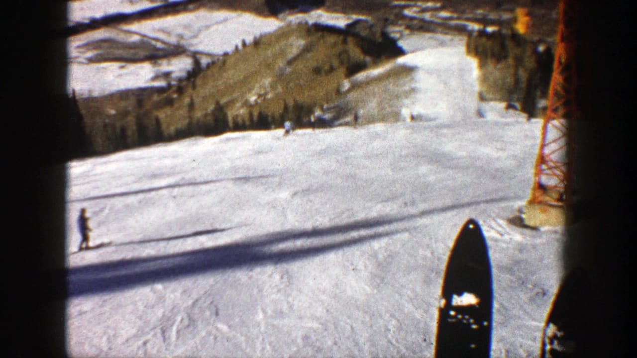 1961 Aspen home-movie frame from a chairlift or elevated POV, showing skis, skiers on a groomed slope, and a red lift tower.