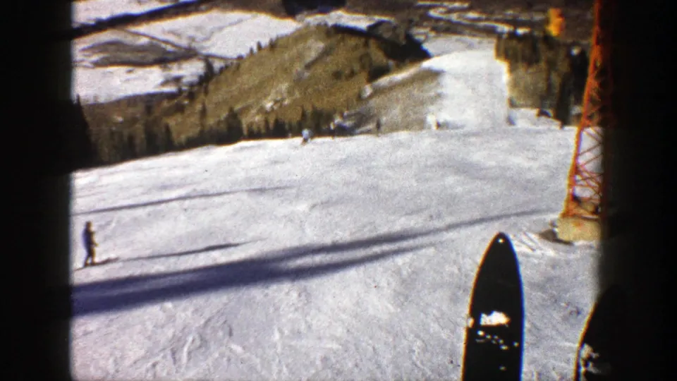 1961 Aspen home-movie frame from a chairlift or elevated POV, showing skis, skiers on a groomed slope, and a red lift tower.