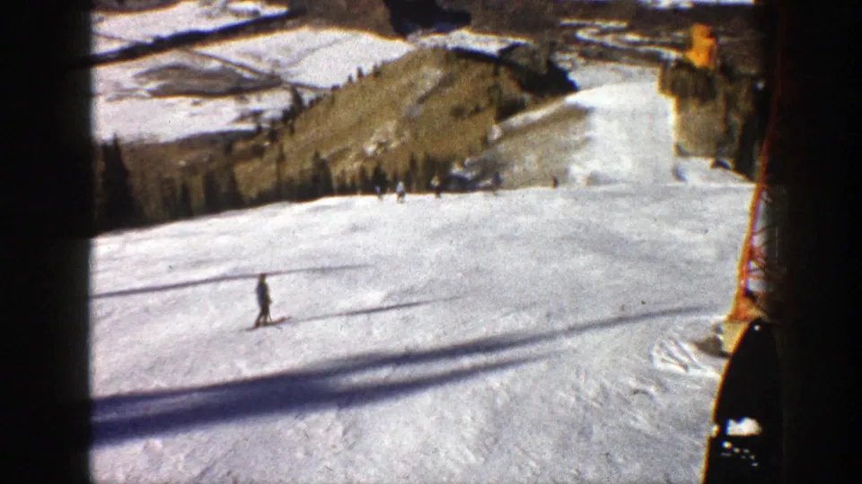 A distant skier moves across the Aspen ski slope in a 1961 8mm frame, with chairlift shadows and a red tower visible.