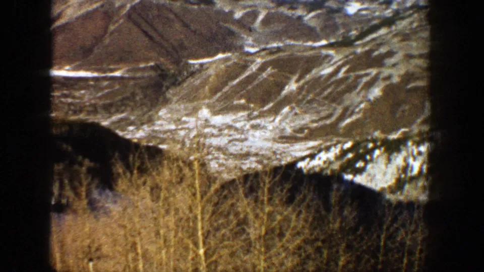 The final 1961 Aspen frame looks away from the ski run toward mountain terrain, patchy snow, and foreground brush.