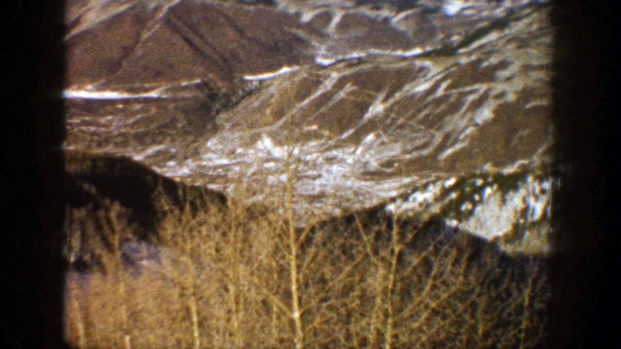1961 Aspen, Colorado home-movie frame showing snowy mountain ridges beyond leafless aspen trees and brush.