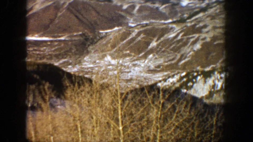 1961 Aspen, Colorado home-movie frame showing snowy mountain ridges beyond leafless aspen trees and brush.