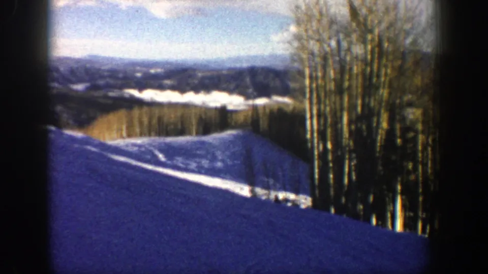 1961 Aspen, Colorado frame showing a snowy foreground slope, a cluster of bare aspen trunks, distant hills, and a pale clouded sky.