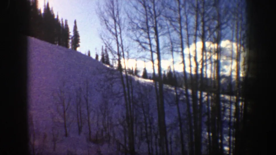 1961 Aspen, Colorado 8mm frame looking past a steep snowy slope toward bare trees, evergreens, clouds, and ridgelines.