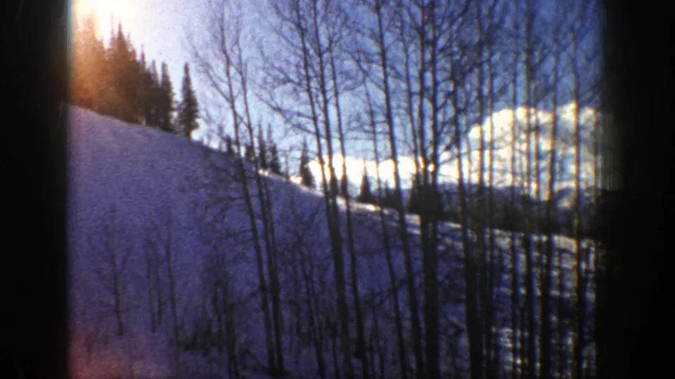 1961 Aspen, Colorado 8mm frame of a snowy hillside, thin aspen trunks, evergreens, clouds, and warm sun flare.