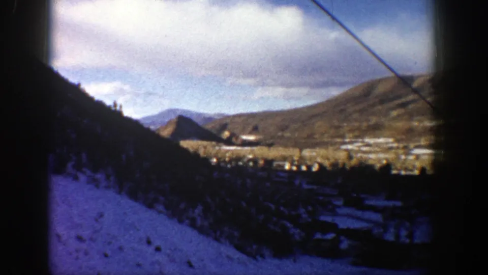 1961 8mm frame from Aspen showing a snowy slope, dense trees, distant valley settlement, and clouded mountain backdrop.
