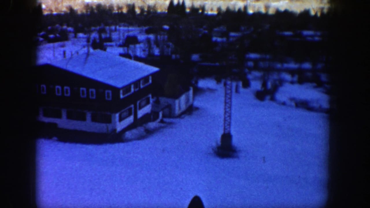 1961 Aspen frame with a lodge roof, tall lift tower, broad snowfield, and wooded background under a blue film tint.