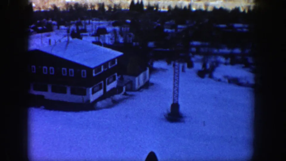 1961 Aspen frame with a lodge roof, tall lift tower, broad snowfield, and wooded background under a blue film tint.