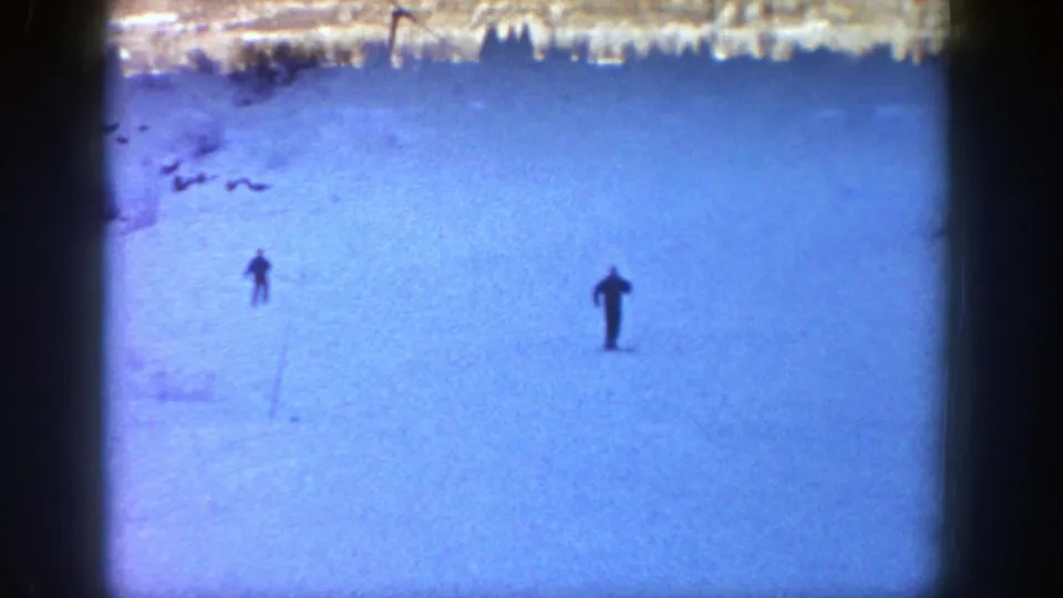 1961 Aspen home-movie frame showing two small silhouetted skiers on a wide snow-covered hill.