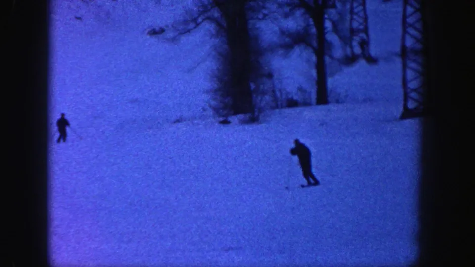Skiers in dark clothing cross a blue-tinted snowy Aspen hillside, with trees and a lift tower visible in the background.