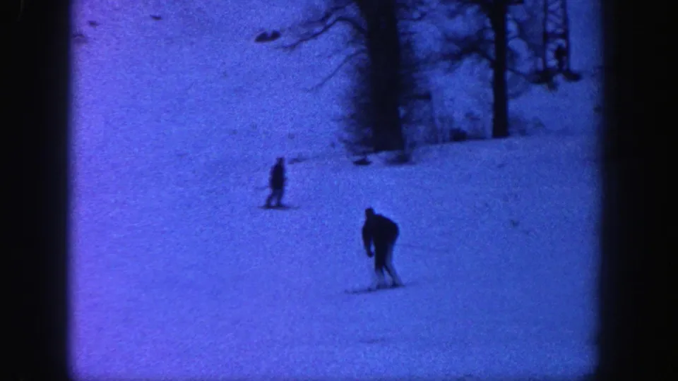 A skier bends forward while descending the Aspen snow slope, with another distant skier uphill in soft 8mm focus.
