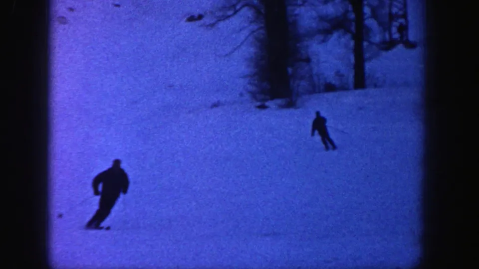 A dark-clad skier crosses the lower part of a blue-tinted Aspen ski slope, with a second skier farther back near the trees.