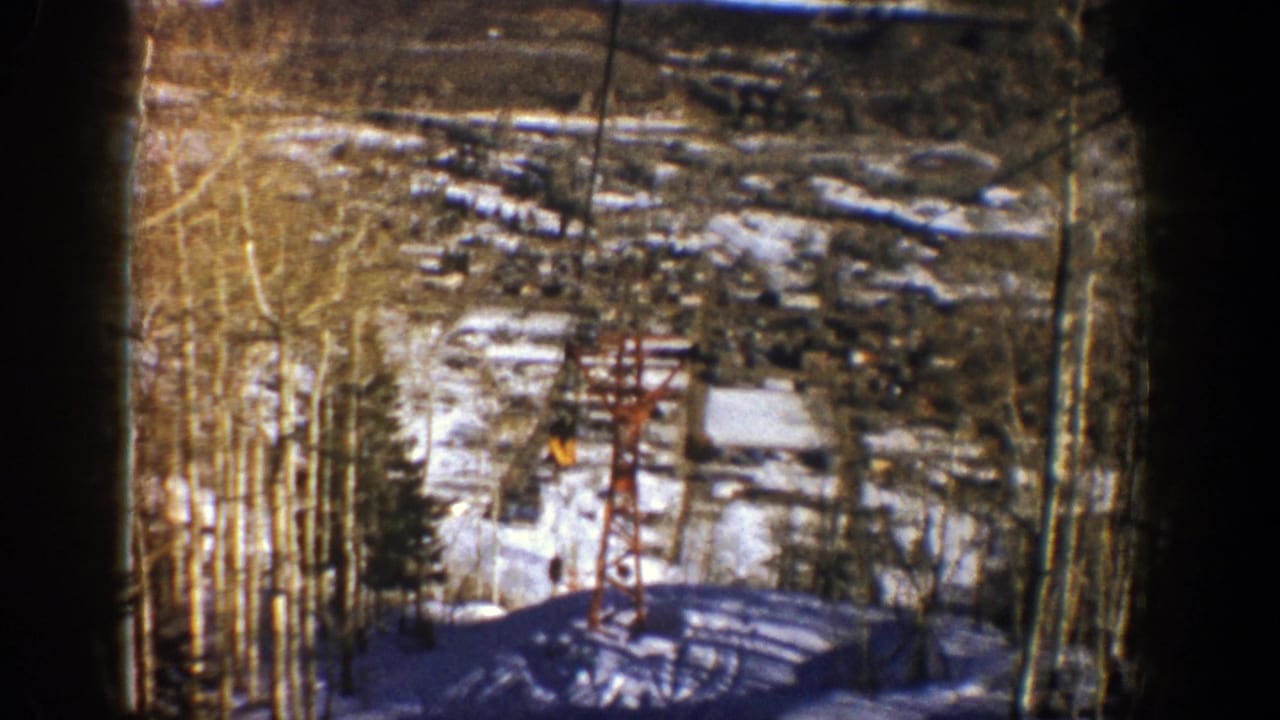 Open chairlift passes a red tower above snow, trees, and distant resort structures.