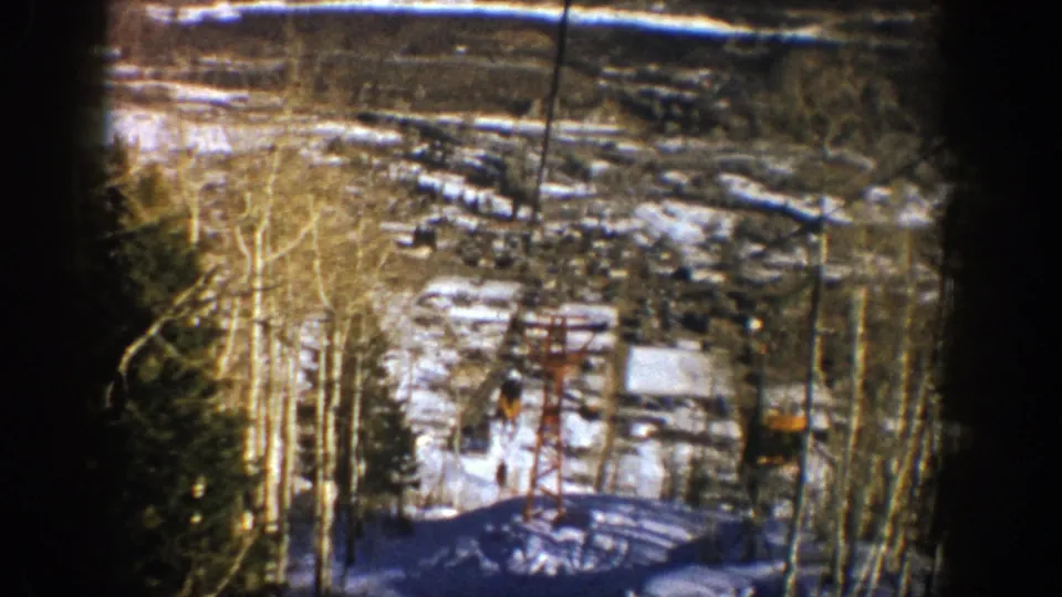 1961 Aspen, Colorado frame showing the chairlift descending past a red tower over a snowy slope with trees and resort buildings beyond.