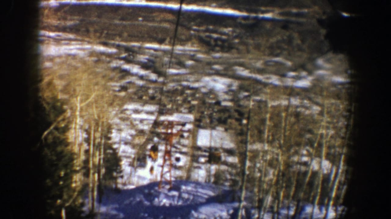 Chairlift cables cross a snowy wooded slope with buildings in the distance.