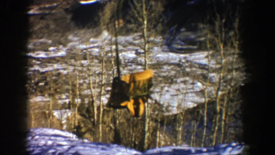 Yellow open chairlift chair descending through bare aspen trees over snowy Aspen terrain in 1961 home-movie footage.