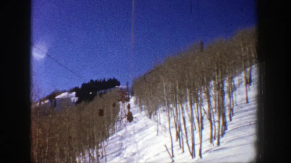 1961 Aspen 8mm frame looking across a bright ski slope, bare aspen trunks, and blue winter sky from the lift route.