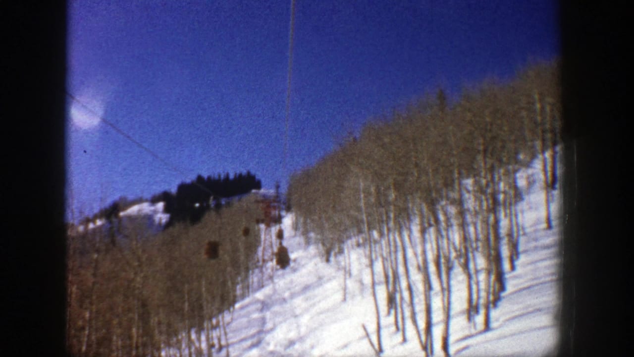 Snowy hillside with bare trees, lift cable, and small brown ski lift cabins under blue sky.